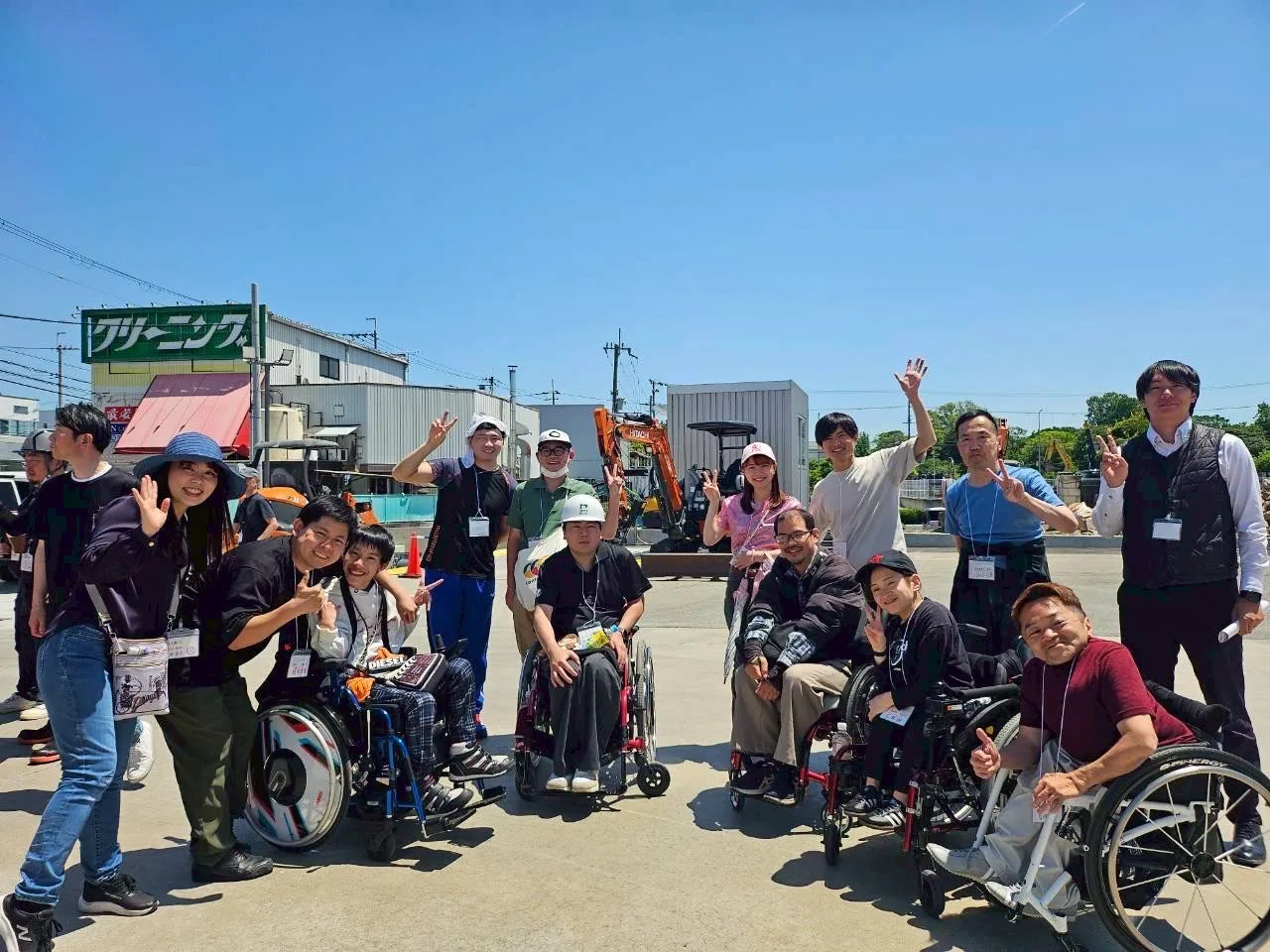 バックホウを背景に車いすユーザーを含む参加者グループが笑顔でピースサインをしている